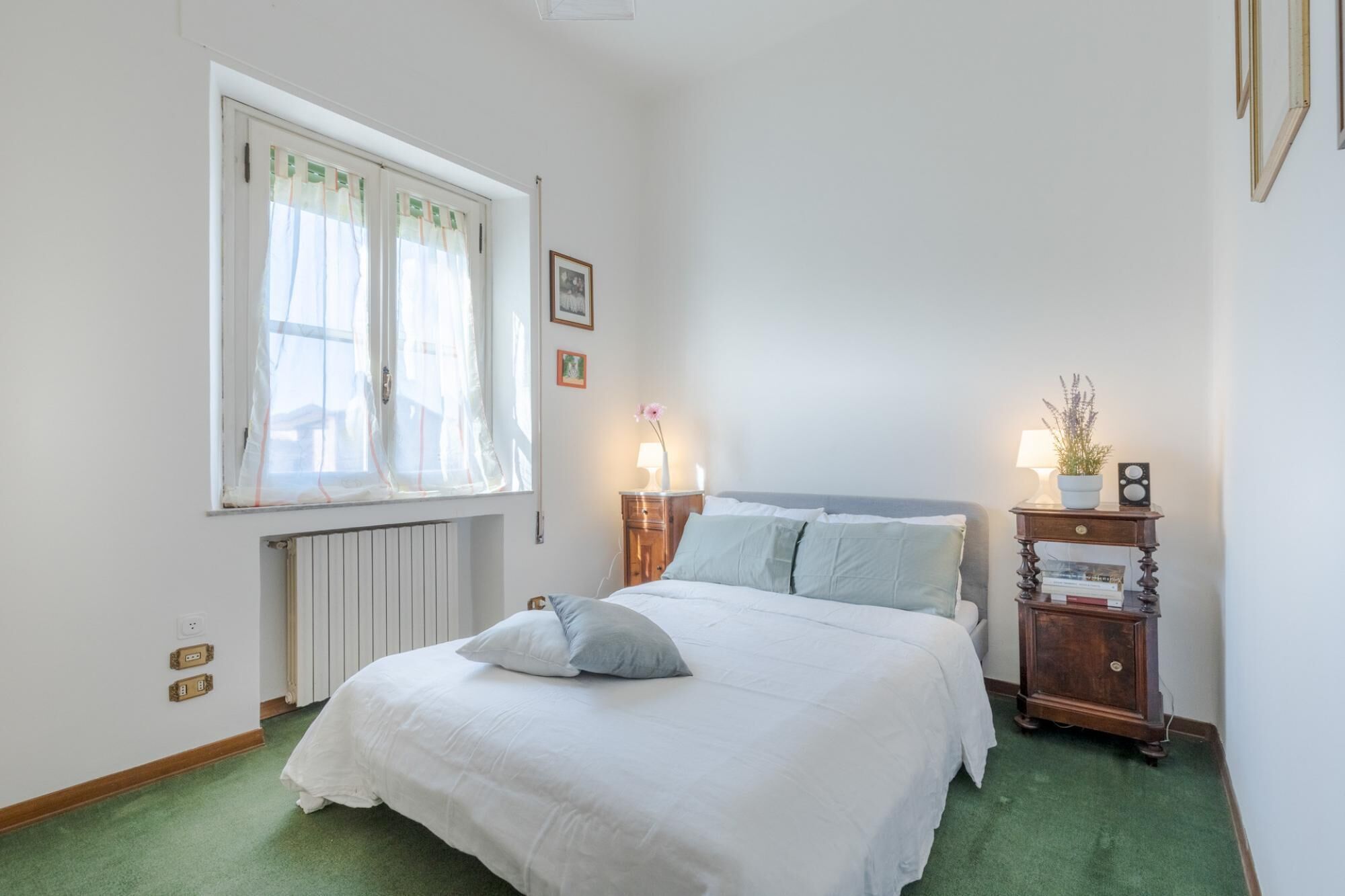 Un dormitorio con cama, mesitas de noche, un radiador, una ventana con cortinas y una alfombra verde.