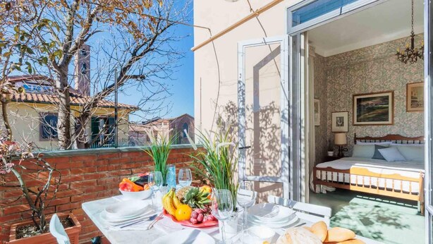 A dining area with a table set for two, a bed with a floral bedspread, and a view of a building with a tiled roof. - La Casa in Centro in Lucca (Lucca)