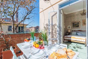 Un coin repas avec une table dressée pour deux personnes, un lit recouvert d’une couvre-lit à motifs floraux et une vue sur un bâtiment dont le toit est recouvert de tuiles.
