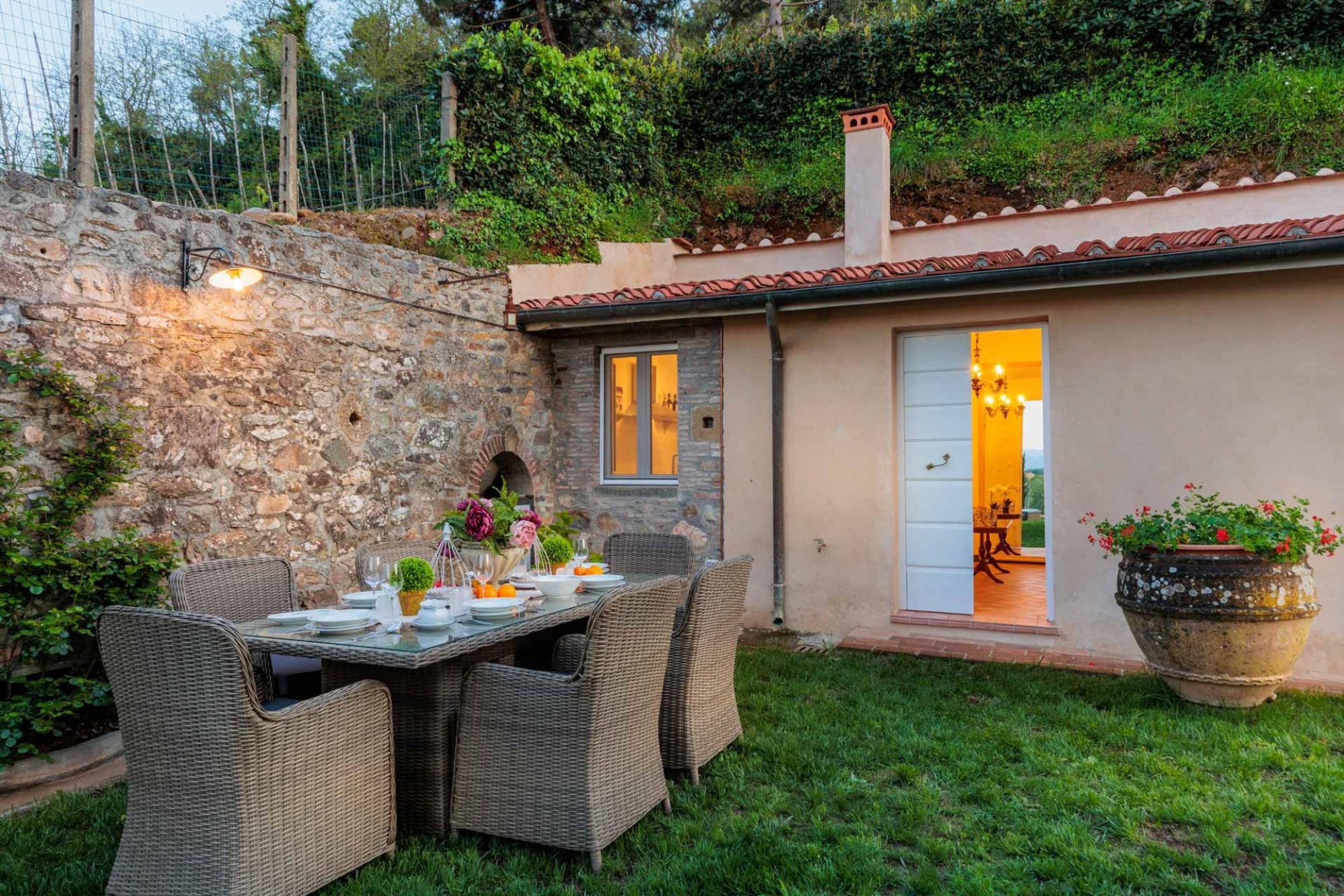 A patio with a table set for dining, surrounded by wicker chairs, a stone wall, and a small building with a tiled roof.