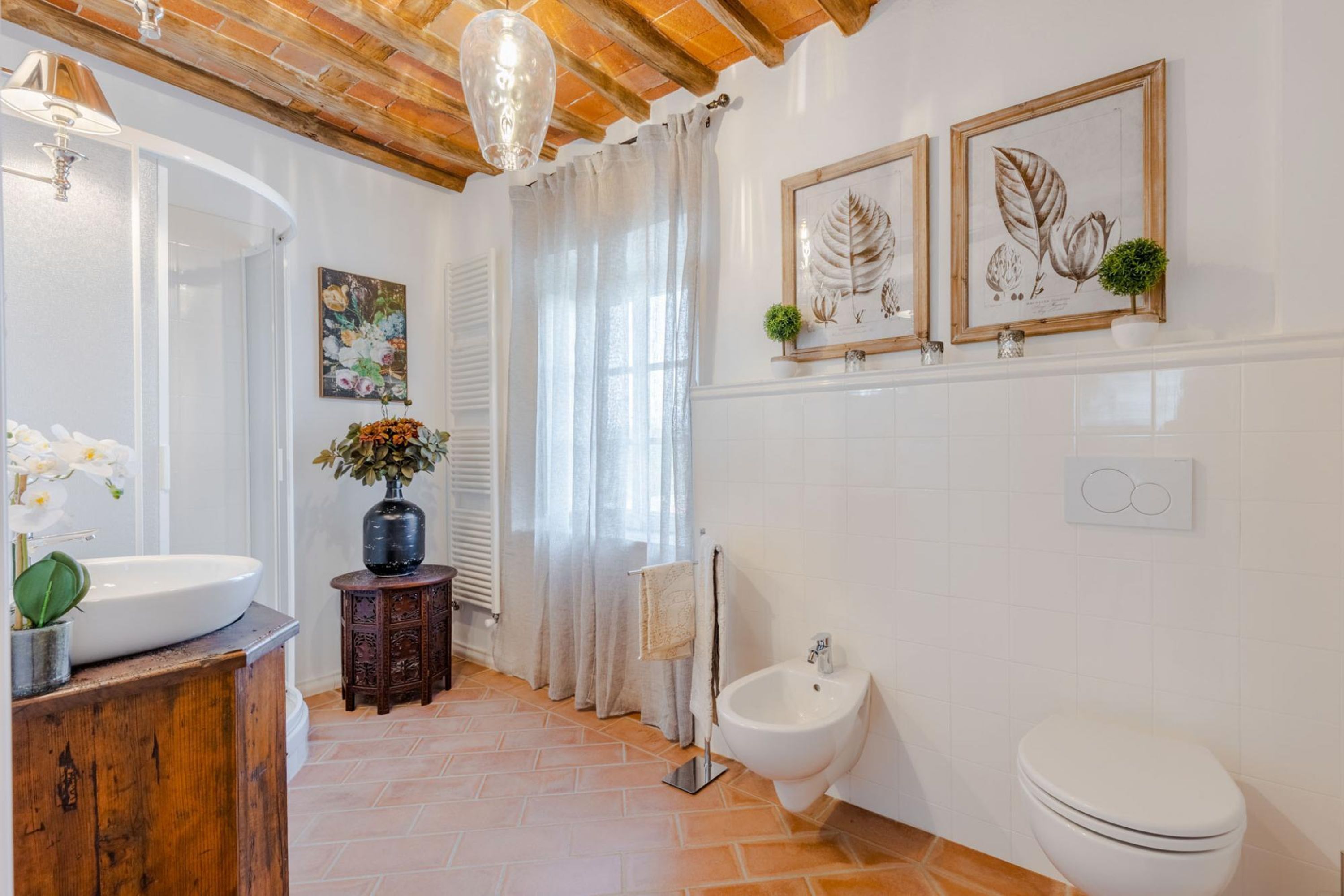 Een badkamer met een witte wastafel, houten wastafelmeubel, een bidet en een wit toilet. Twee ingelijste botanische prints hangen aan de muur, er staat een kleine pot met een plantje en een vaas met bloemen op het wastafelmeubel.