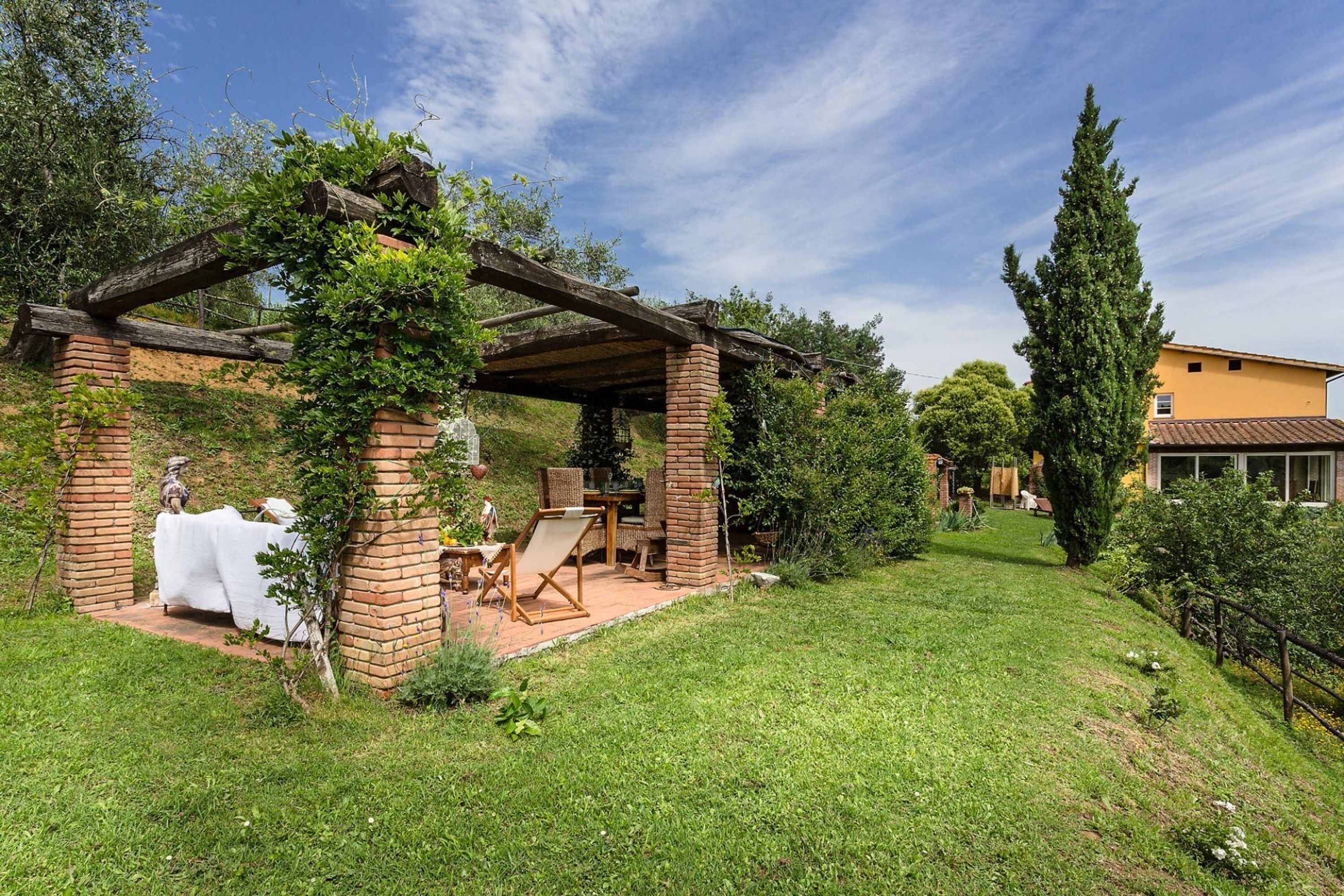 Ein überdachter Außenbereich mit Ziegelstützen und einer Pergola, umgeben von Grünpflanzen und blauem Himmel.