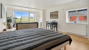 Un dormitorio con una cama grande, una zona de comedor con una mesa y sillas, un ventanal con vistas a un edificio rojo y una planta en maceta.