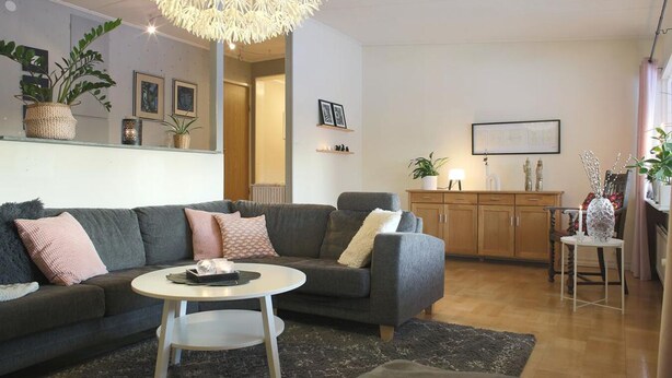 Un salon moderne avec un canapé gris, une table basse ronde et un lustre. On y trouve également un meuble en bois et une petite table ornée d’objets décoratifs.