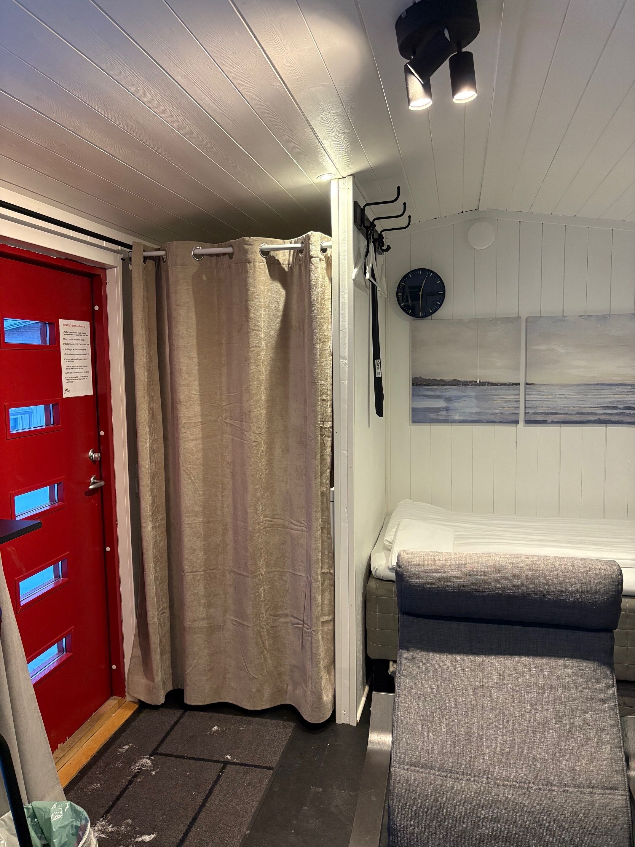 A room with a red door, beige curtains, a bed with a gray headboard, a clock on the wall, and a small window.