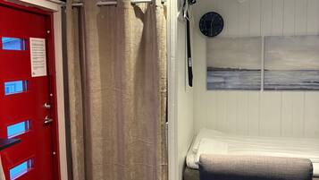 A room with a red door, beige curtains, a bed with a gray headboard, a clock on the wall, and a small window.