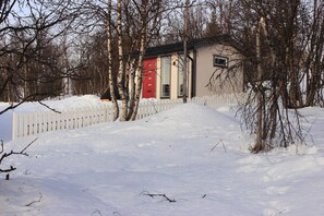 A snow-covered landscape with a white picket fence, bare trees, and a small building with a red door. - Aurora Cabin (Kiruna)