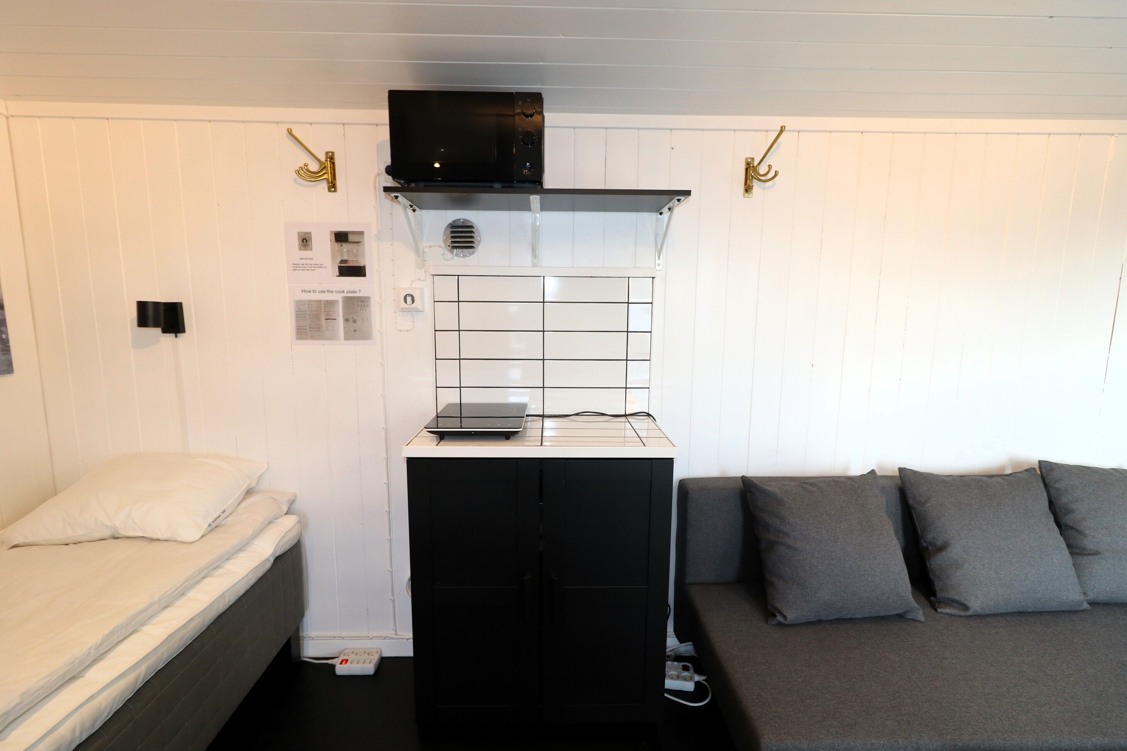 A compact room with a bed, a sofa, a small kitchenette with a microwave, and a wall-mounted shelf.