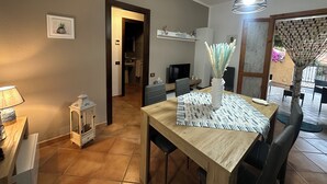 Un coin repas avec une table en bois, des chaises, un vase contenant des fleurs séchées et un téléviseur en arrière-plan.