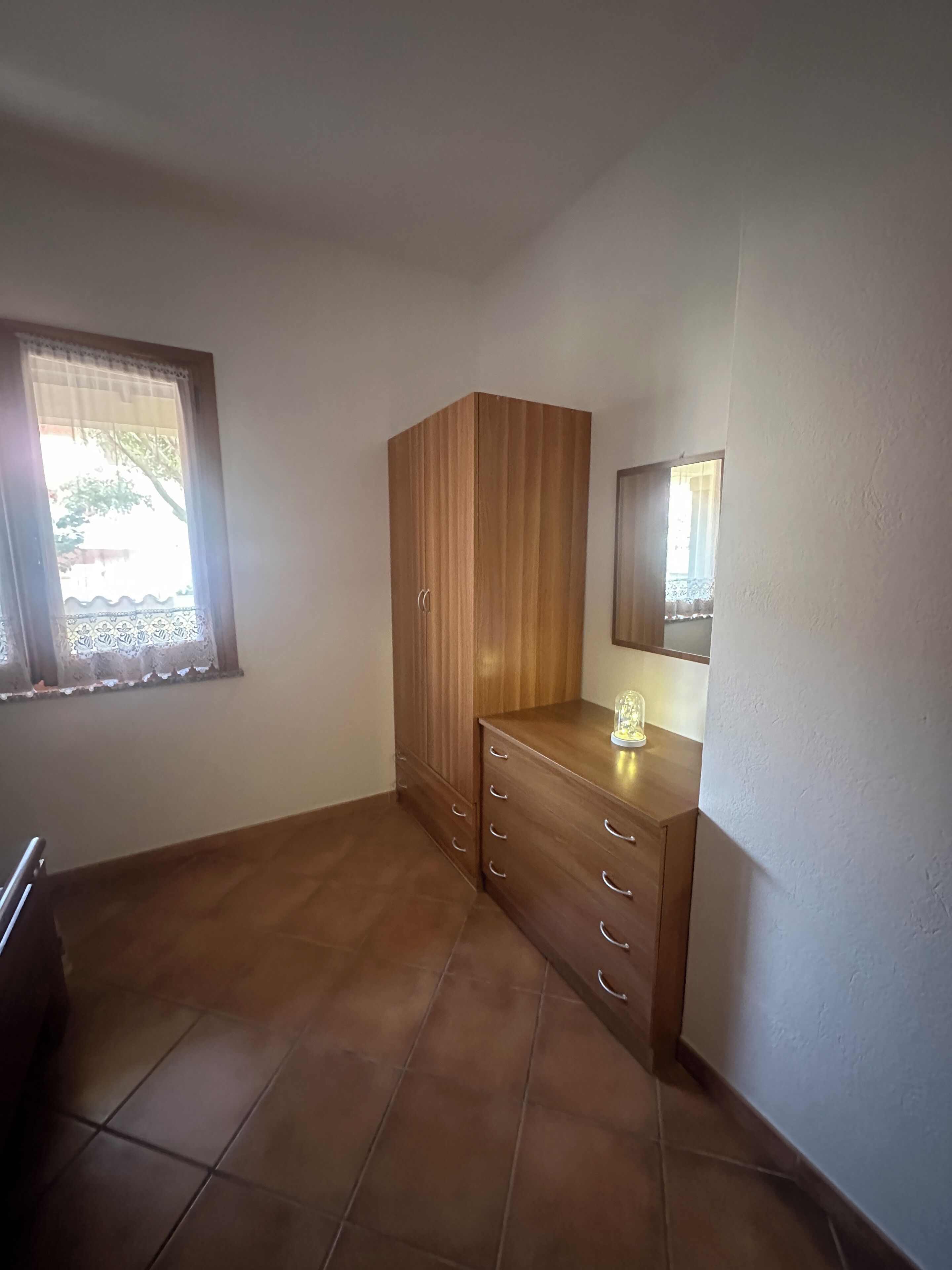 A room with a wooden wardrobe, a small table with a lamp, and a window with lace curtains.