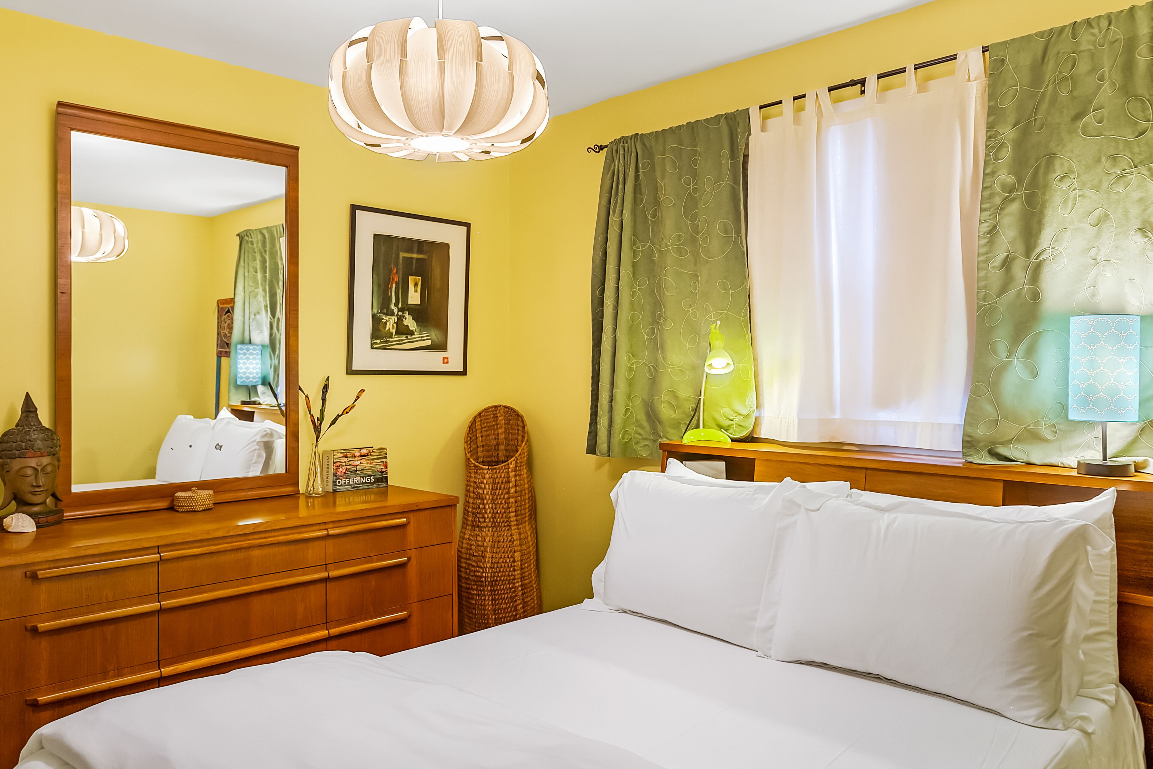 A hotel room with a bed, wooden dresser, mirror, artwork, and a window with curtains.