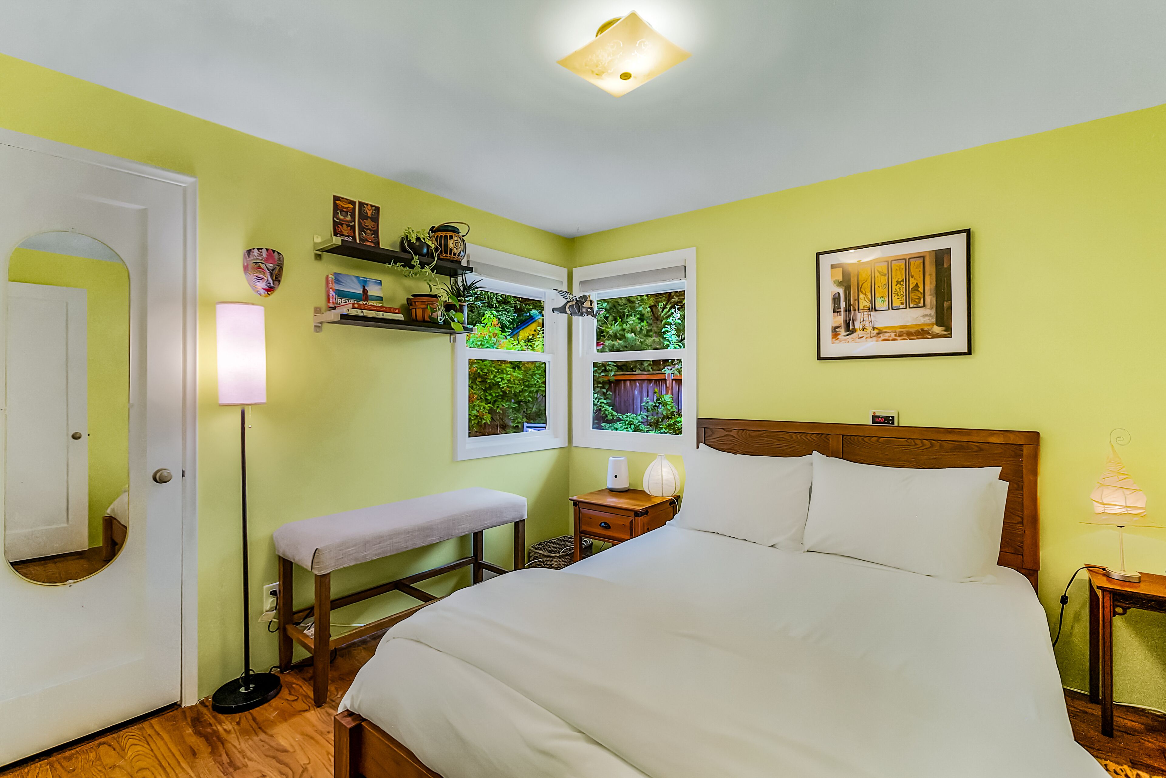 Un dormitorio con cama, mesitas de noche, un banco, una puerta y ventanas con vistas a zonas verdes.