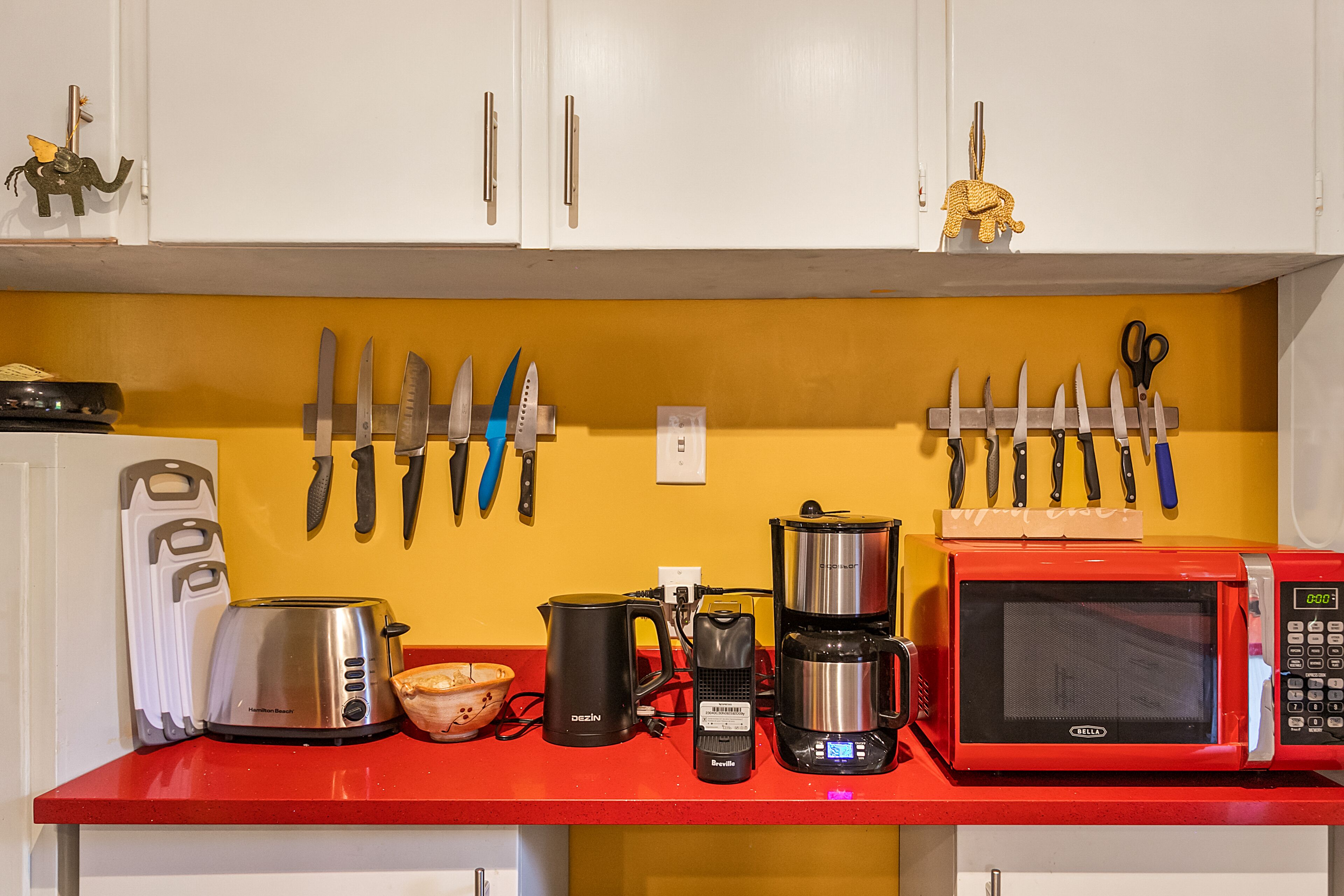 Une cuisine avec des meubles blancs, un plan de travail rouge et divers appareils électroménagers, notamment un grille-pain, une machine à café et un four à micro-ondes.