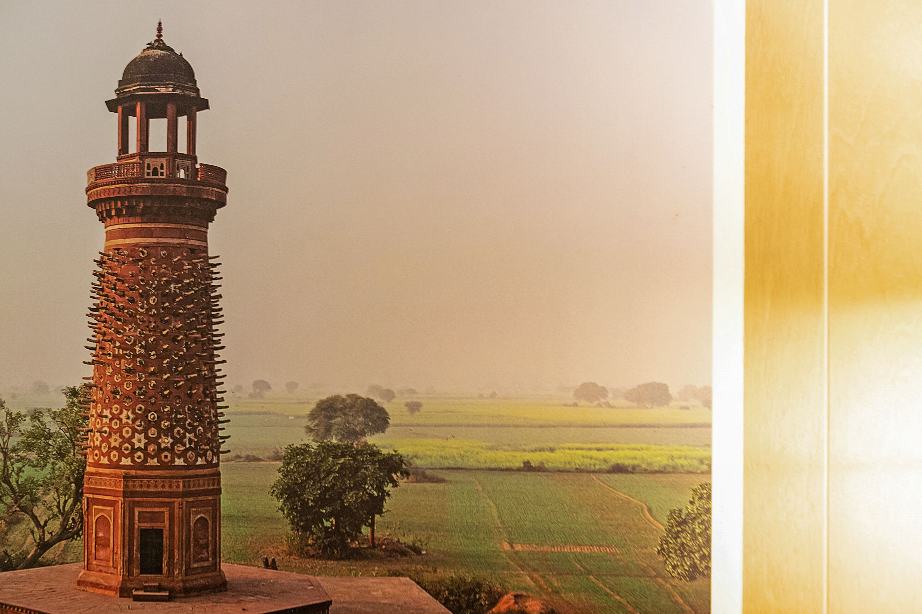 A tower with intricate designs and a dome, surrounded by fields and trees.