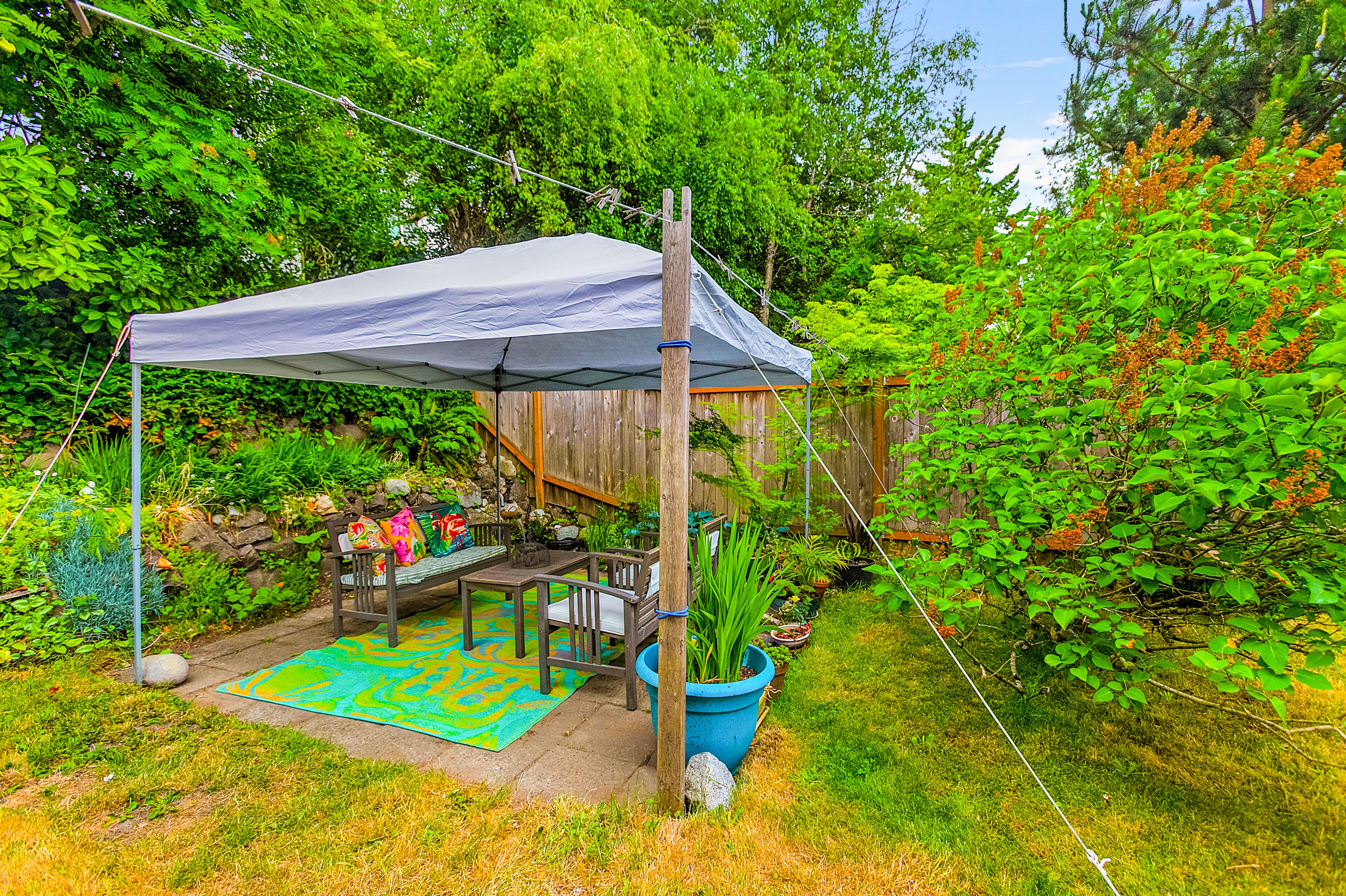 A covered outdoor seating area with a wooden deck, a stone wall, and lush greenery.