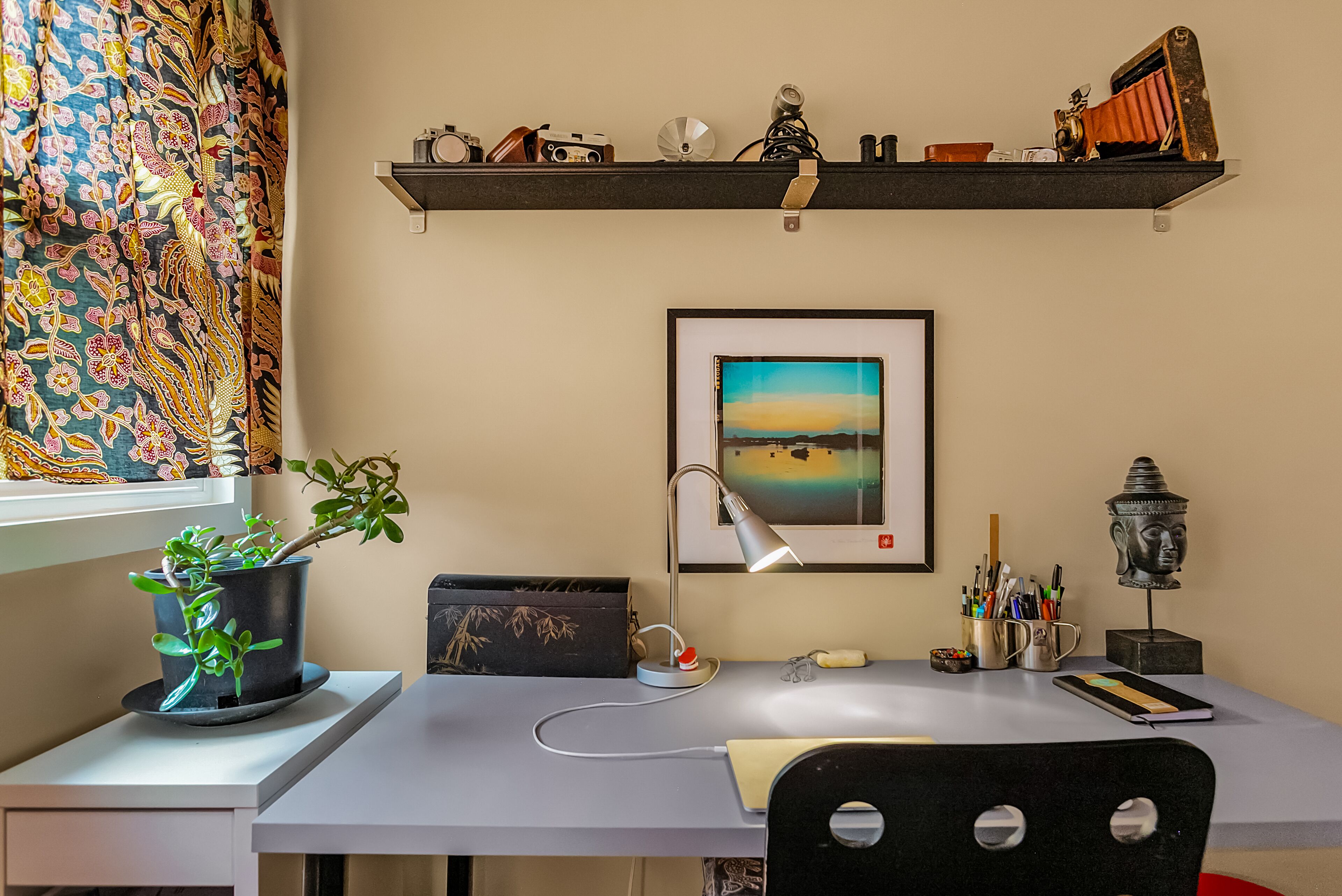 Un bureau avec un ordinateur portable, un cadre photo, une lampe de bureau, une plante en pot et une statue de Bouddha.