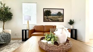 En stue med en brun lædersofa, et rundt vævet sofabord med planter, et indrammet landskabsmaleri på væggen og en potteplante ved vinduet.