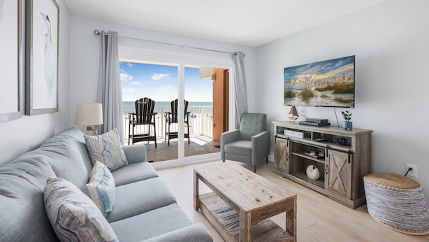 En moderne stue med en grå sofa, et træbord og et fjernsyn på et træskab. Der er en balkon med udsigt til stranden.