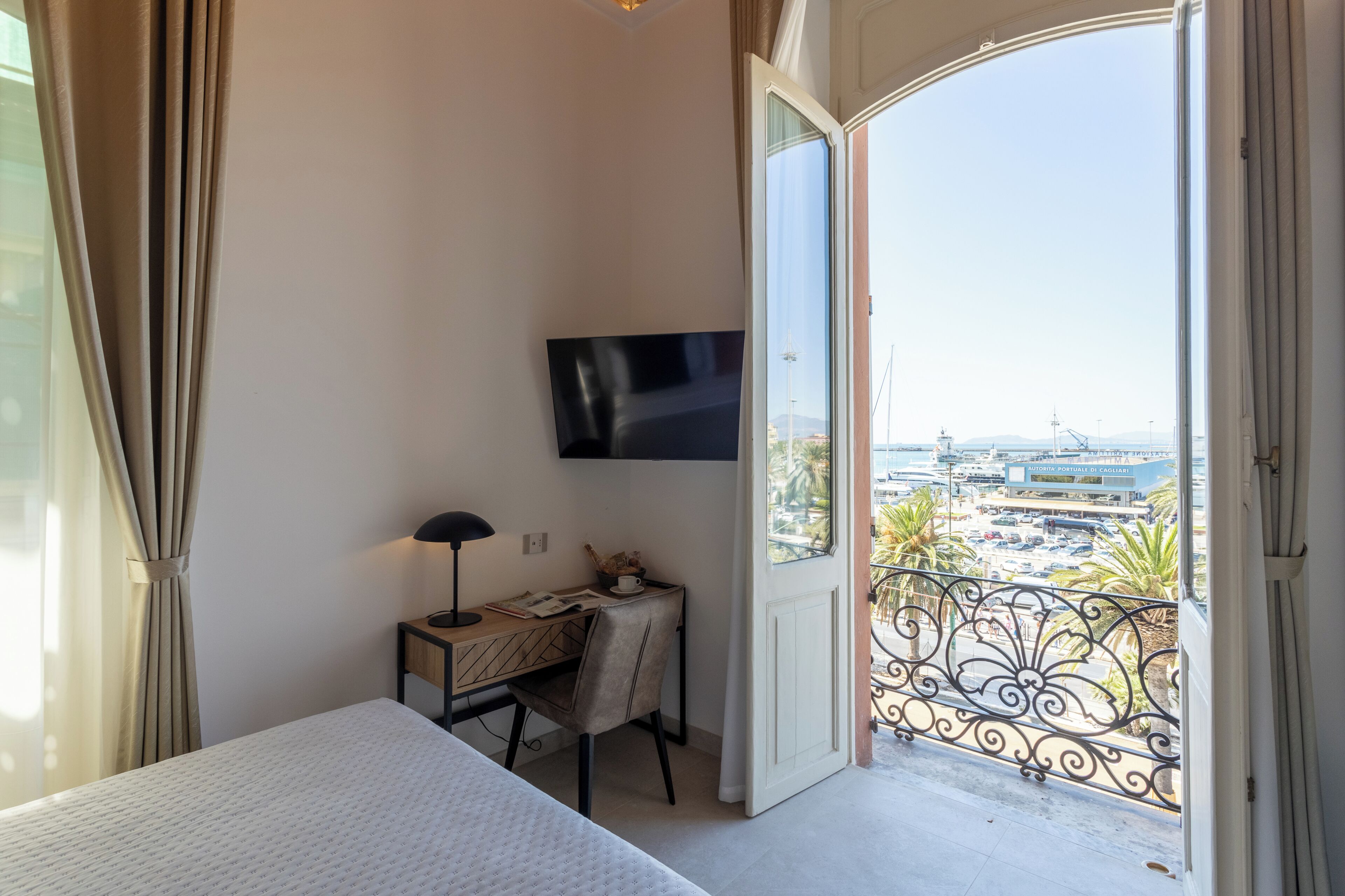 A hotel room with a bed, a desk with a lamp, a TV, and a balcony with a view of the city.