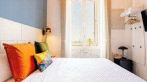 A hotel room with a bed, pillows, a nightstand, a lamp, a TV, and a view of the outside. - Magnini Rooms (Cagliari)