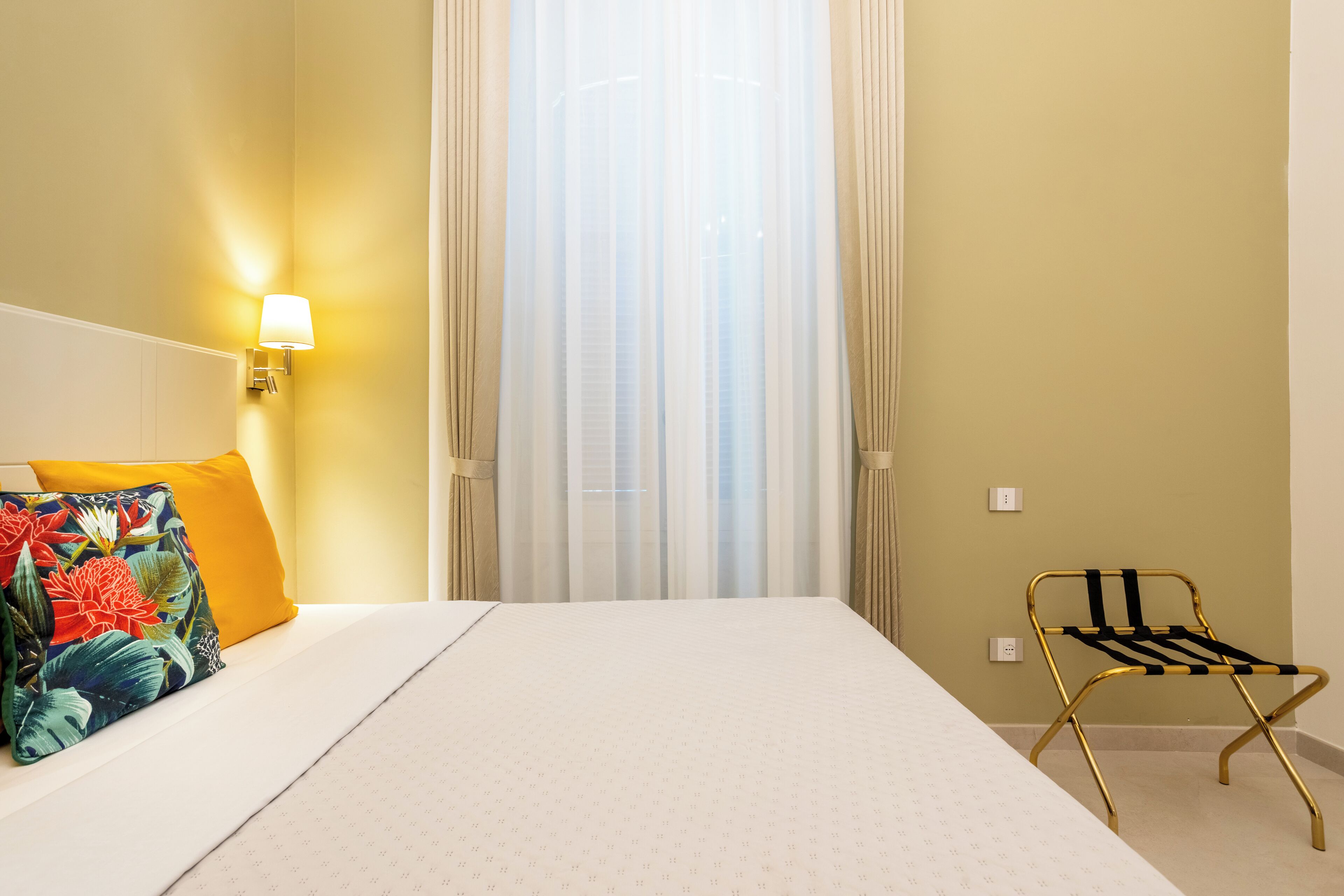 Ein Hotelzimmer mit Bett, Nachttisch, Lampe, Stuhl und Fenster mit durchsichtigen Vorhängen.