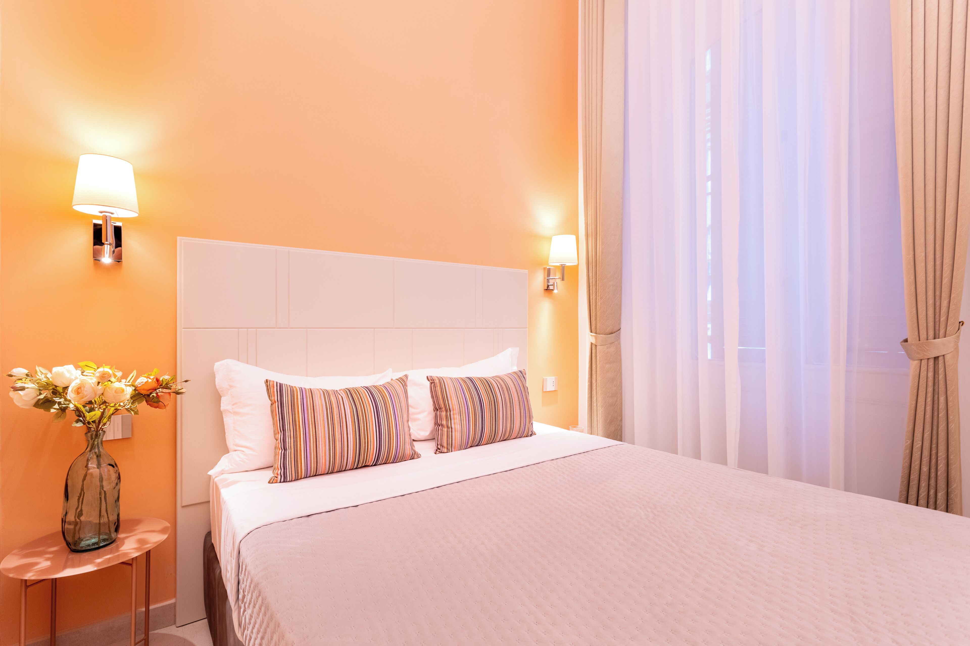 Une chambre d’hôtel avec un lit, deux lampes fixées au mur, un vase de fleurs et des rideaux en voile.