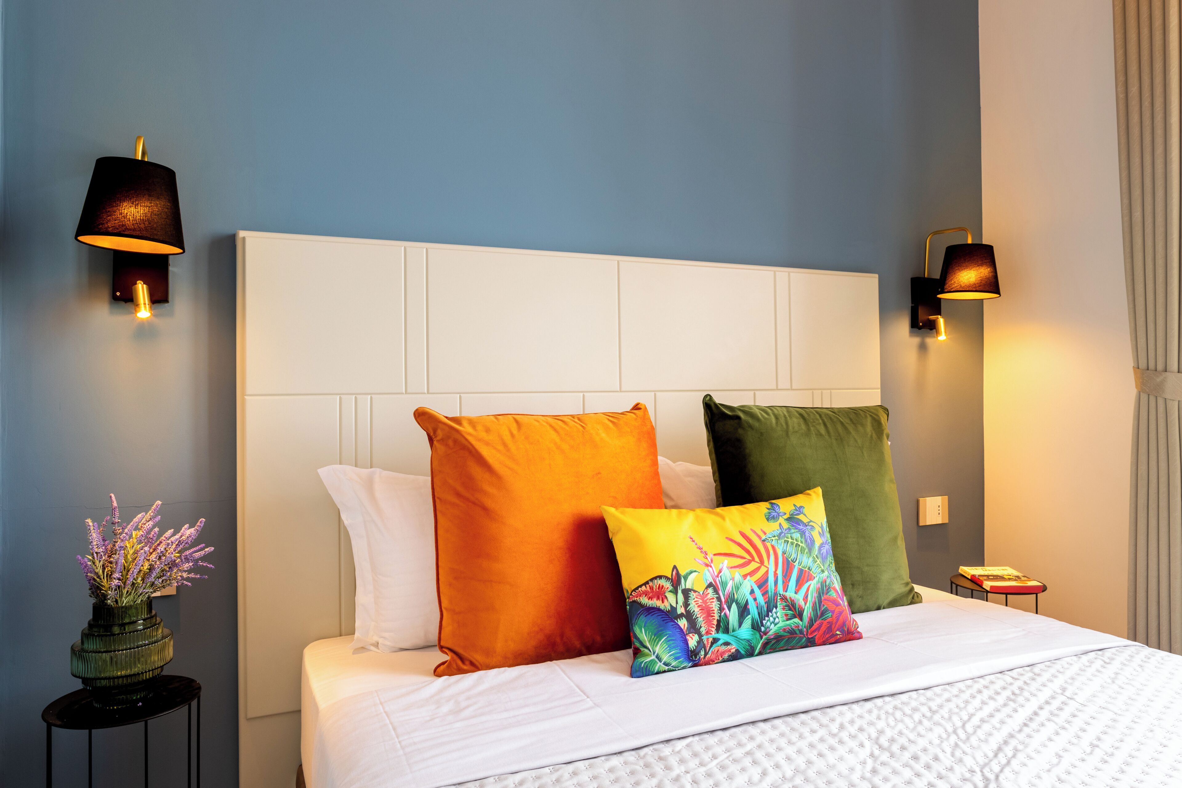 A hotel room with a white headboard, a bed with white bedding, and colorful pillows. There are two wall-mounted lamps on either side of the bed.