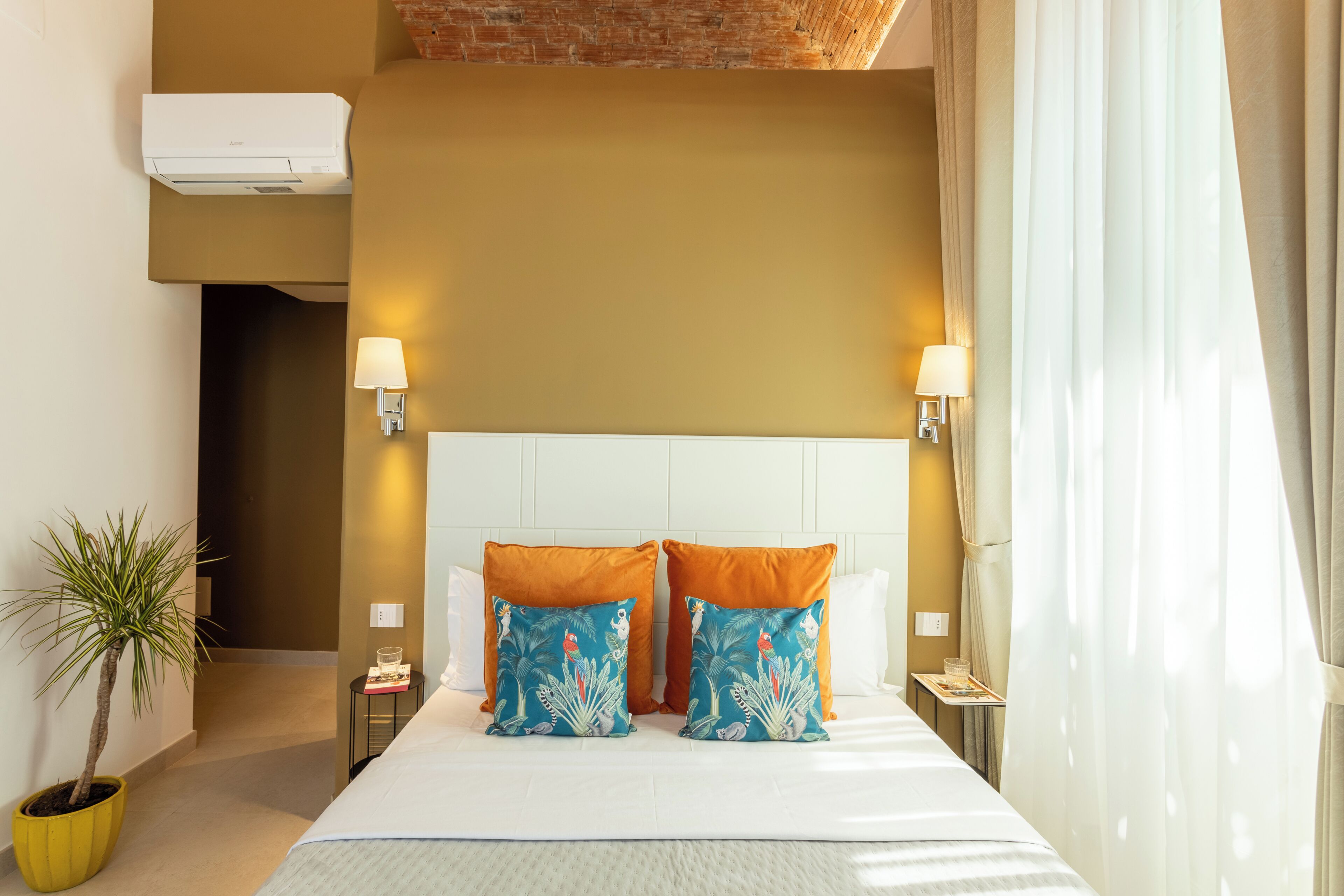 A hotel room with a large bed, two wall-mounted lamps, a potted plant, and a brick wall.