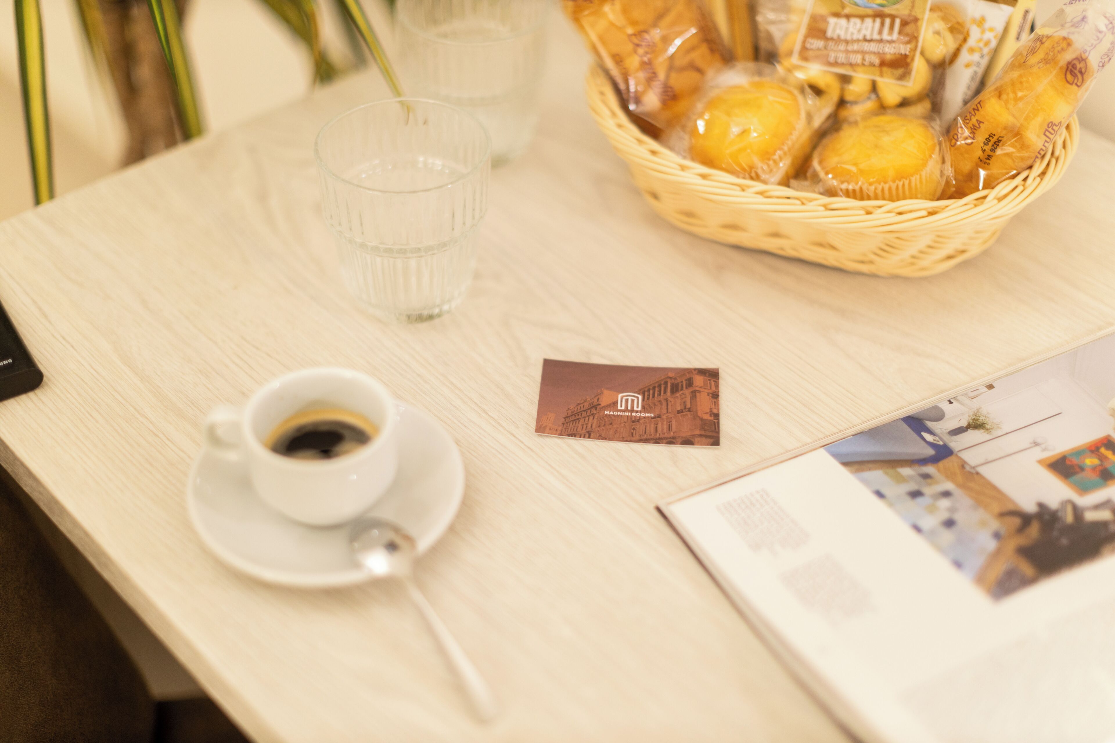 A table with a cup of coffee, a small card, a basket of pastries, and a brochure.