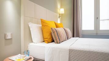Een hotelkamer met een bed, kussens, een nachtkastje met een glas water en een raam met gordijnen.