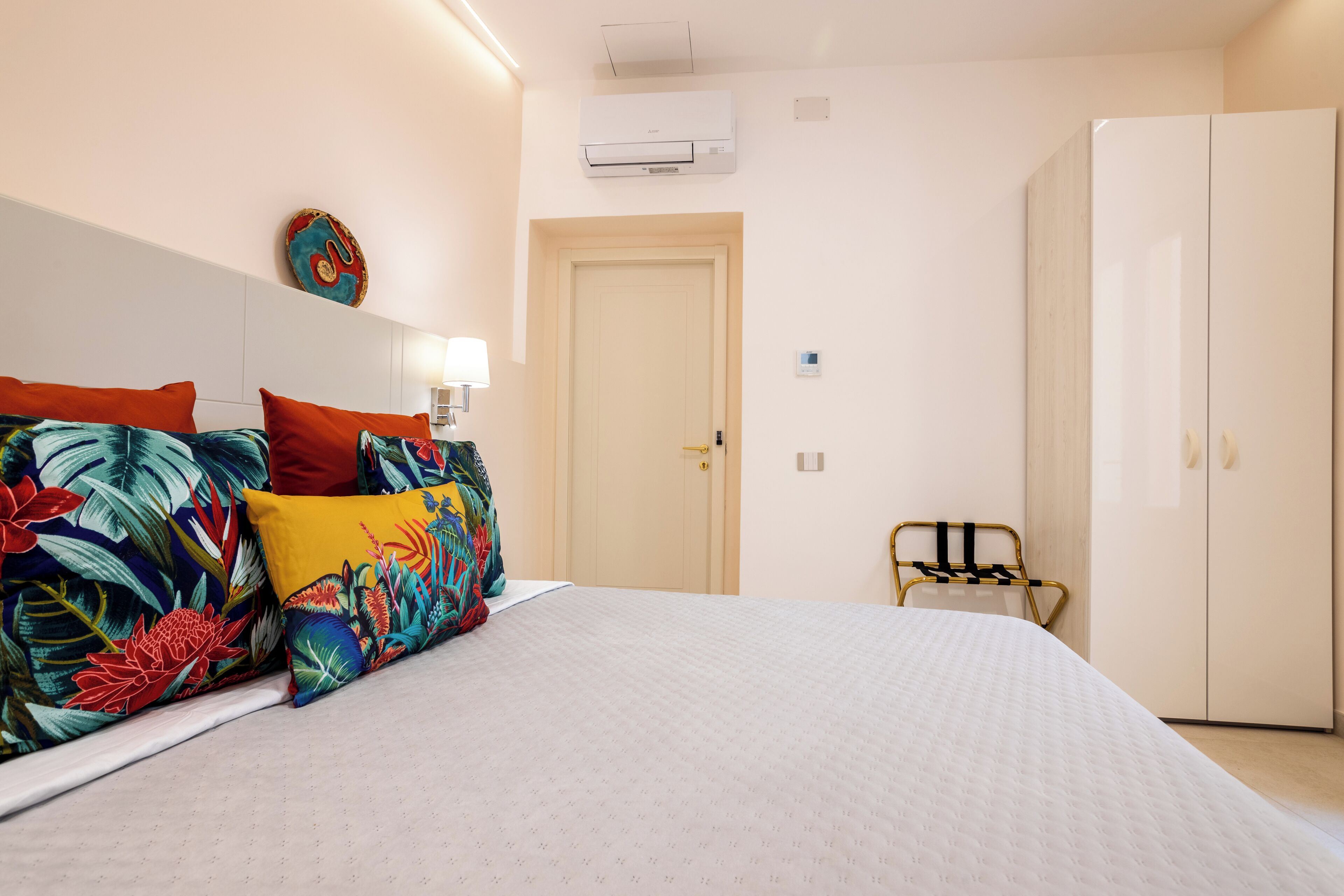 A hotel room with a bed, a wardrobe, a bedside table, a lamp, and an air conditioning unit.