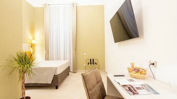 A hotel room with a bed, a desk with a chair, a television, a plant, and a basket of snacks.