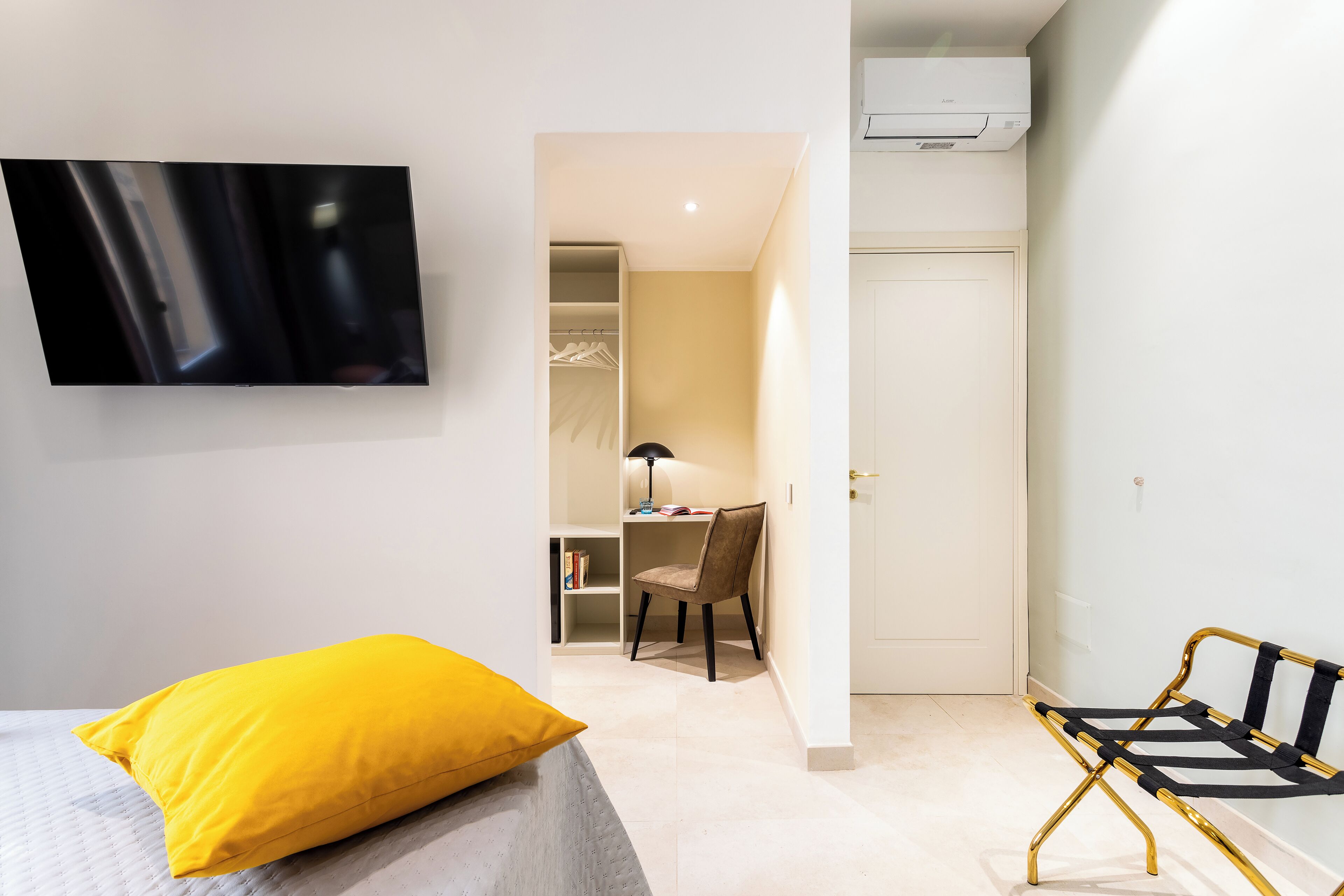 A bedroom with a bed, a yellow pillow, a desk with a chair, and a TV mounted on the wall.