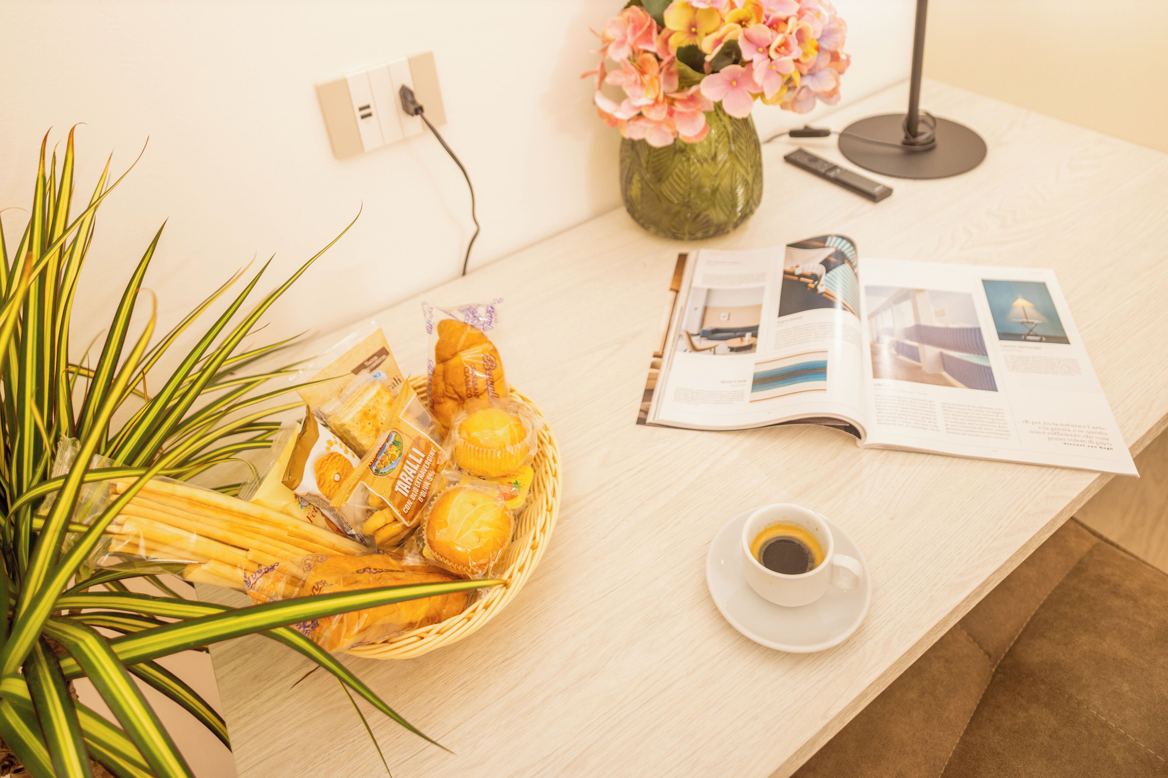 A breakfast setup with a basket of pastries, a cup of coffee, a vase of flowers, and a magazine on a wooden table.