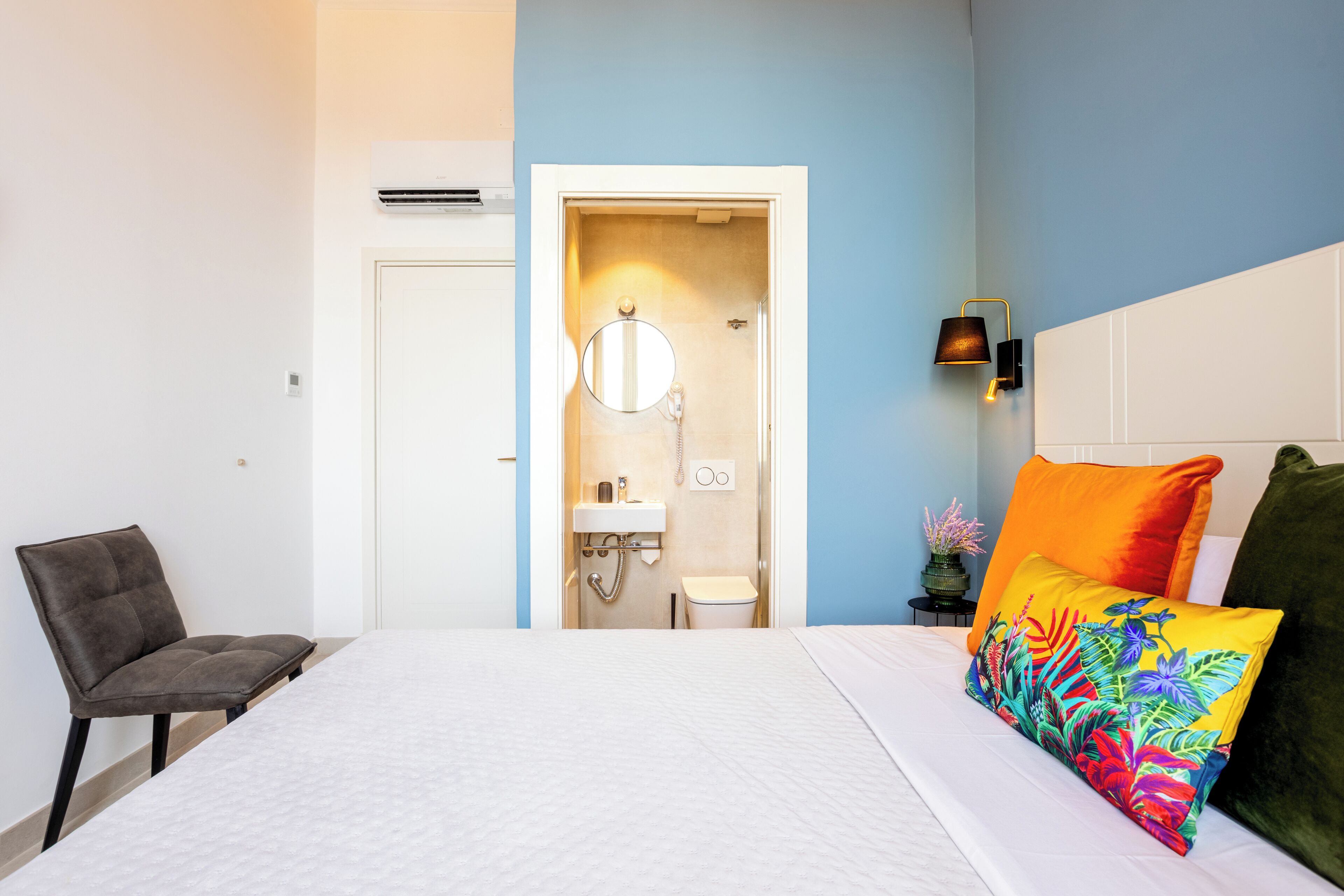 A bedroom with a bed, a chair, a bedside table with a lamp, and a view into the bathroom.