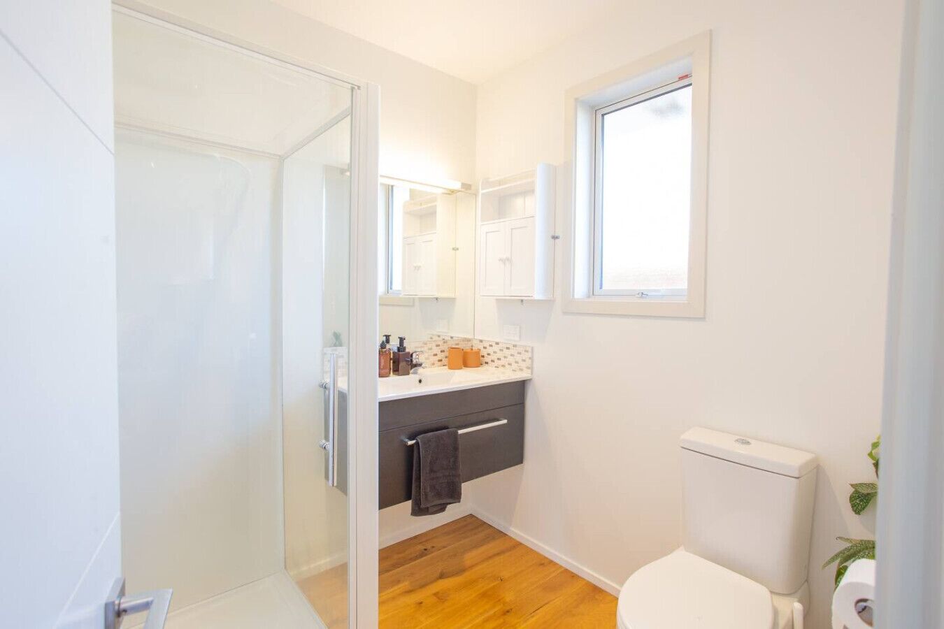 Une petite salle de bain dotée d’une porte de douche en verre, de toilettes blanches, d’un lavabo avec un pare-éclaboussures carrelé et d’une fenêtre située au-dessus du miroir.