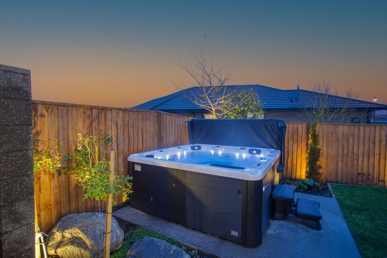 A hot tub in a backyard with a wooden fence, a small garden, and a house in the background.