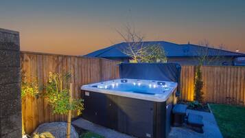 A hot tub in a backyard with a wooden fence, a small garden, and a house in the background.