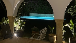 Un área de patio al aire libre con piscina, rodeada de plantas en macetas y una mesa de madera.