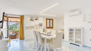 Una cocina moderna con armarios blancos, una zona de comedor con una mesa y sillas blancas, y vistas a un jardín a través de una ventana.