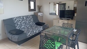 Un salon avec un canapé, une table à manger à plateau en verre et des chaises. On y trouve également un téléviseur posé sur un meuble, un réfrigérateur et un miroir accroché au mur.