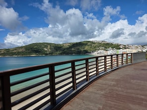 A coastal walkway with a railing, a clear view of the sea, and a hillside with buildings. - Aconchegante Apartamento na Prainha (Arraial do Cabo)