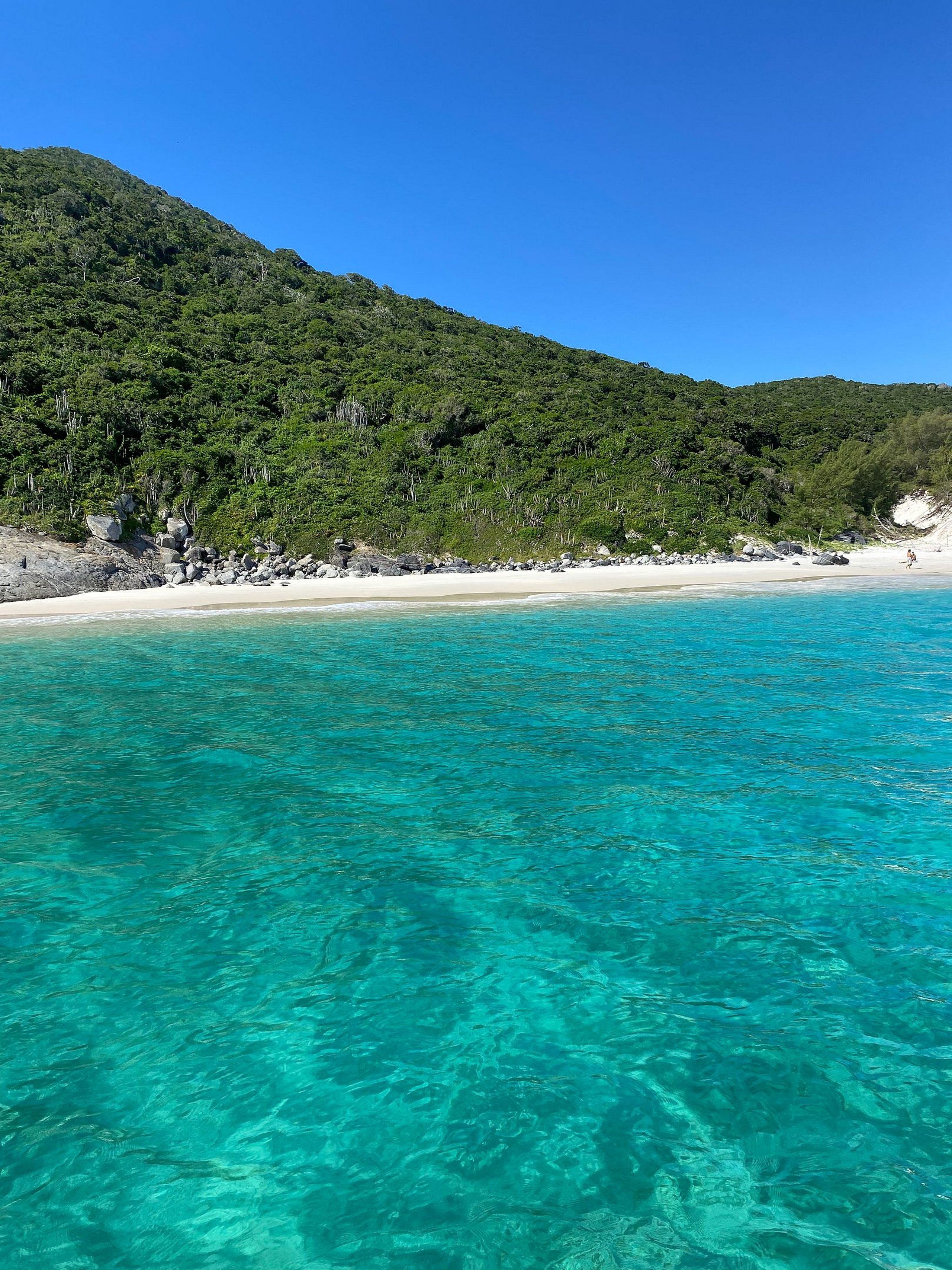 Uma praia com águas turquesa cristalinas, areal arenoso e uma colina arborizada ao fundo.