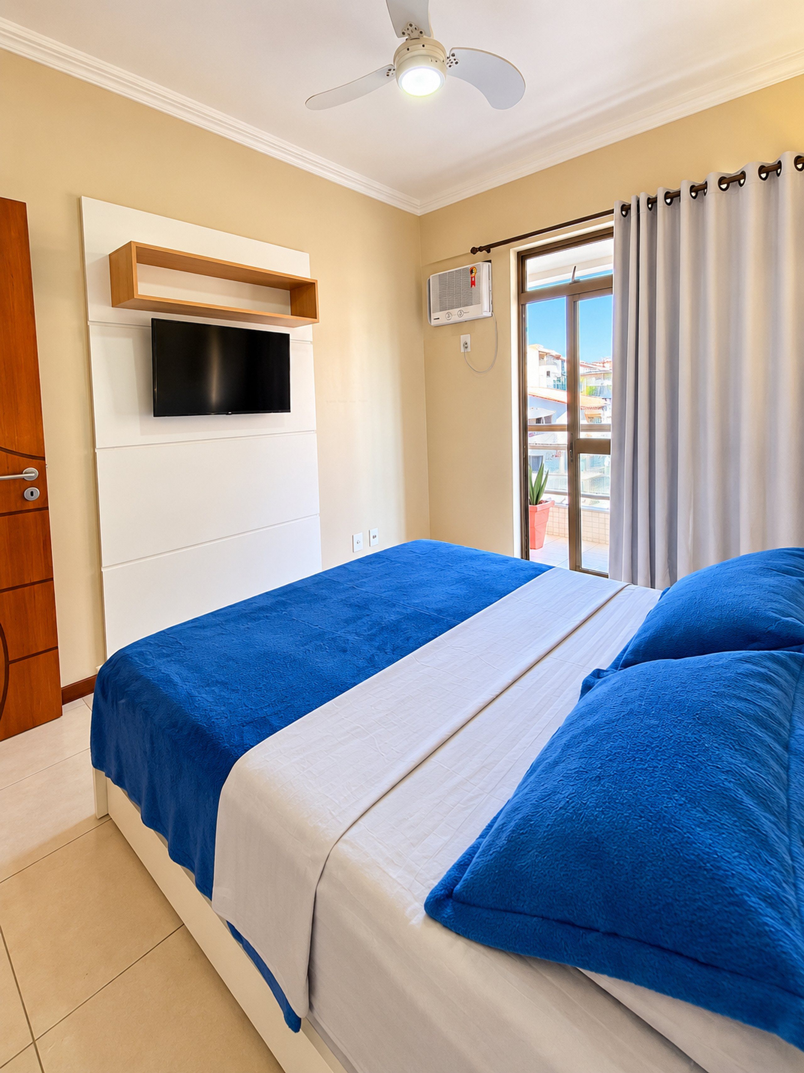 A hotel room with a bed, a television mounted on the wall, a ceiling fan, and a window with curtains.