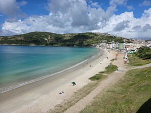 A coastal beach with a clear view of the sea, a sandy shore, and a hillside with buildings.