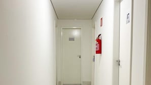 A narrow hallway with white walls and a marble floor, leading to a door labeled "Bancomero." A fire extinguisher is mounted on the wall to the right. - Hotel Lara 10 Curitiba (Curitiba)