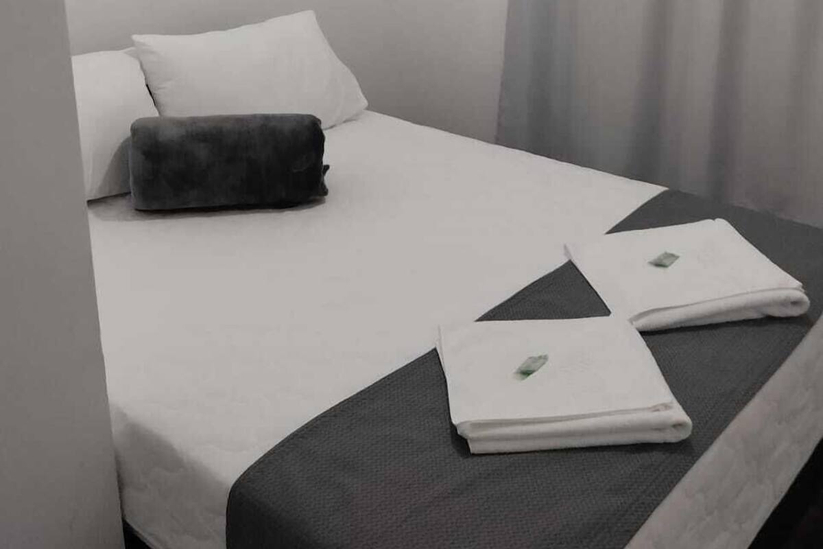 A neatly made bed with white and dark grey bedding, two folded towels, and a grey pillow.