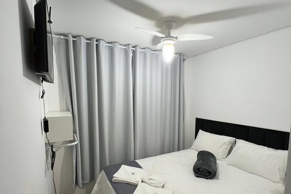 A hotel room with a bed, a ceiling fan, a TV mounted on the wall, and a window with curtains.