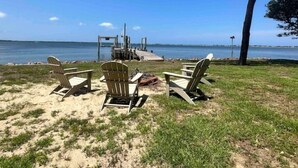 Una playa con tres sillas Adirondack mirando al agua, un muelle de madera y un árbol.
