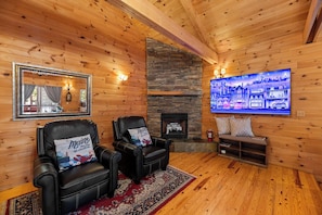 A cozy cabin interior with a stone fireplace, leather chairs, a large mirror, and a television displaying a cityscape.